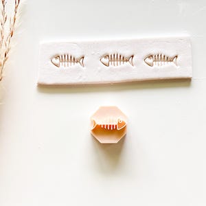 May include: A light pink rectangular mold with three fish skeleton impressions and a light pink octagonal stamp with a fish skeleton design. The stamp and mold are on a white surface.
