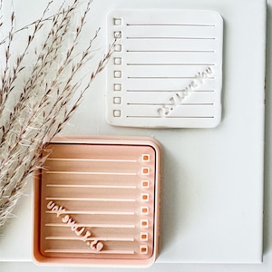 May include: A white square notepad with horizontal lines and checkboxes, featuring the embossed text "I Love You," alongside a peach-colored square tray with matching lines, checkboxes, and the embossed text "I Love You" in a different orientation.