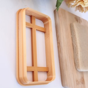 May include: A light brown plastic cookie cutter shaped like a window with a grid pattern. The cutter is on a white surface.