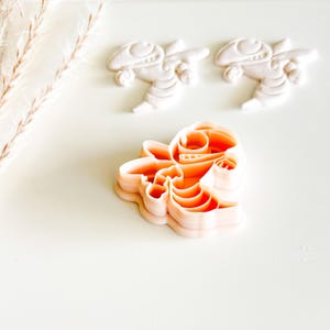 May include: An orange plastic cookie cutter in the shape of a cartoon character is displayed with two white, molded figures of the same character. The cutter is in the foreground, with the white figures in the background.