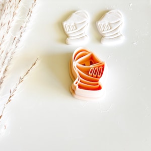 May include: Three knight helmet cookie cutters. Two are white, and one is orange. The cookie cutters are on a white surface. The knight helmet design is detailed with lines and shapes.