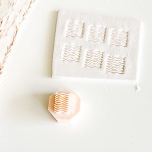 May include: A light peach-colored, octagonal stamp with a wavy pattern, next to a white rectangular mold with the same wavy design. The mold has six recessed areas. The stamp is likely for creating decorative patterns.