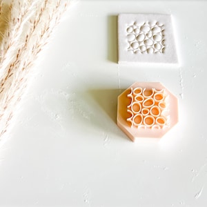 May include: Two craft tools are displayed. One is a white square with a pattern of raised, irregular circles. The other is a pink, octagonal stamp with a similar pattern of raised circles. The tools are on a white surface.
