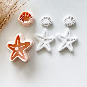 May include: A collection of seashell and starfish-shaped cookie cutters. The cutters are in shades of pink and white. The starfish cutters have five points. The seashell cutters have a detailed design. The cutters are arranged on a white surface.