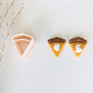 May include: Two miniature pumpkin pie slices with white frosting, and a pink plastic pie cutter.