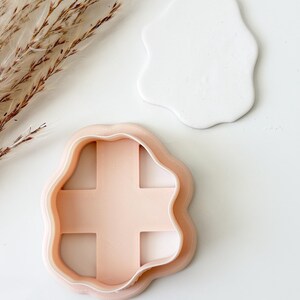 May include: A light peach-colored plastic cookie cutter with an irregular, wavy outline and a cross-shaped interior structure, positioned next to a white, irregular, wavy-shaped piece of clay that has been cut out by the tool.