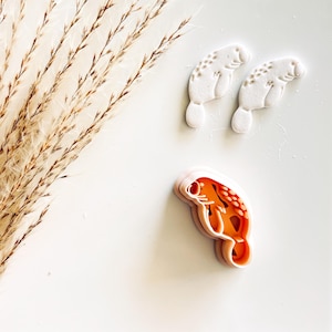 May include: Three manatee-shaped cookie cutters. Two are white, with brown dot patterns. The third is orange and pink, showing the manatee's interior design. Dried wheat stalks are in the upper left corner, all on a white surface.