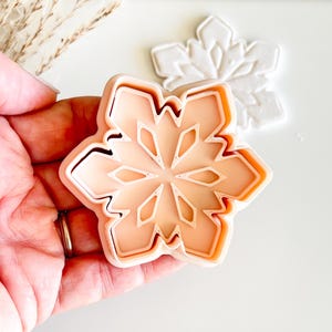 May include: A snowflake-shaped cookie cutter in a light peach color, held in a hand. The cutter has a detailed design with geometric shapes. A white snowflake shape is visible in the background. The cutter is likely used for baking or crafting.