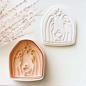 May include: A peach-colored plastic cookie cutter and a white embossed cookie impression featuring a nativity scene with two figures standing on either side of a central manger under a star, all enclosed within an arched frame design.