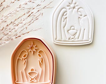Può includere: Un tagliabiscotti in plastica color pesca e un'impronta di biscotto bianca raffigurante una scena della natività con due figure in piedi ai lati di una mangiatoia centrale sotto una stella, il tutto racchiuso in una cornice ad arco.