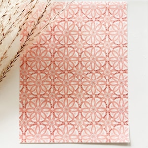 May include: A rectangular sheet of paper with a repeating floral pattern in shades of pink and white. The design features stylized flowers and dotted circles. Dried ornamental grass is in the upper left corner.