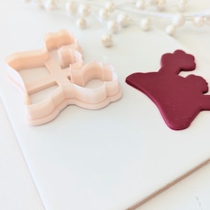 May include: A close-up of two cookie cutters, one in the shape of a crown and the other in the shape of a dress. The crown cutter is made of red fondant and the dress cutter is made of white plastic.