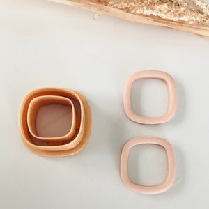May include: Set of three square-shaped cookie cutters. The largest cutter is a light brown color, the medium cutter is a slightly darker brown, and the smallest cutter is a light pink color.