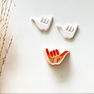 May include: Three white and orange hand-shaped cookie cutters, arranged on a white surface. Two are solid white, while the third has a red and orange interior. The hand gesture is a symbol of the shaka sign, often associated with surfing and the Aloha spirit.