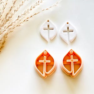 May include: Four earring cutters shaped like a leaf with a cross cut-out. Two are white, and two are orange with a light orange outline. The cutters are on a white surface with dried grass in the background.