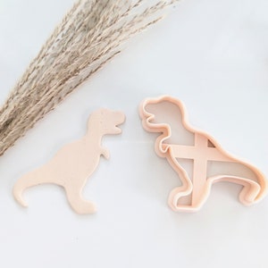 May include: A set of two dinosaur cookie cutters, one solid and one with a cut-out design. The cutters are made of a light pink plastic.