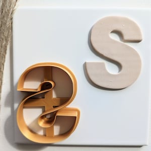 May include: Two cookie cutters in the shape of the letter "S". One is gold and the other is beige.