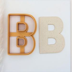May include: A gold plastic cookie cutter in the shape of the letter B, next to a white ceramic letter B.