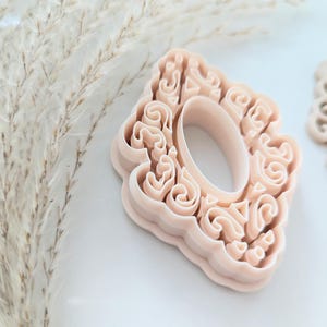 May include: A light pink plastic cookie cutter shaped like a decorative oval frame with intricate swirls and details.