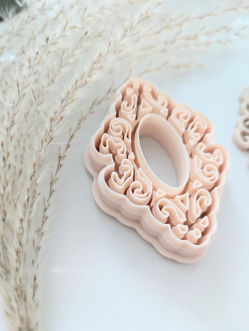 May include: A light pink plastic cookie cutter shaped like a decorative oval frame with intricate swirls and details.