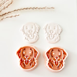 May include: Four dog-shaped cookie cutters. Two are light pink, and two are white. The dog design features a smiling face with a tongue sticking out. The cutters are likely for making dog-shaped cookies or treats.