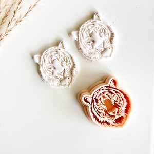 May include: Three tiger head-shaped cookie cutters. Two are white, and one is a light orange color. The cutters are on a white surface. The tiger heads have detailed facial features and ear shapes.