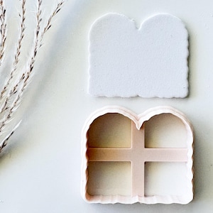 May include: A white arch-shaped cookie cutter with a scalloped edge and a cross-shaped internal divider, accompanied by a matching white arch-shaped piece of cut dough, both featuring a smooth, matte finish and a consistent, uniform shape.