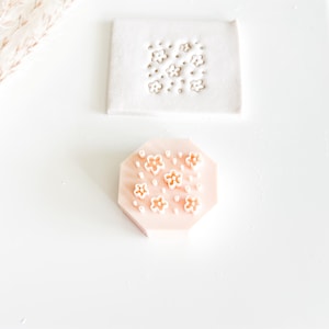 May include: A light pink, octagonal clay stamp with a floral design. The stamp features small, raised flower shapes and dots. A white clay square with the same floral pattern is above the stamp. The background is white.