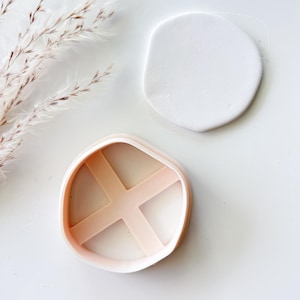 May include: A peach-colored plastic cookie cutter with an irregular, rounded shape and an internal cross-shaped support structure, positioned next to a smooth, white, circular piece of clay or dough on a plain white surface.