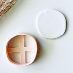 May include: A peach-colored circular cookie cutter featuring a cross-shaped internal support structure, positioned next to a smooth, flat, circular white clay disc that has been cut out using the tool.