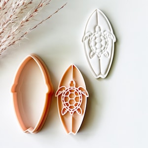 May include: A peach-colored surfboard-shaped cookie cutter with a matching white stamp insert featuring a sea turtle design. The set includes an open outline cutter and a detailed press tool that imprints the turtle shape onto dough.