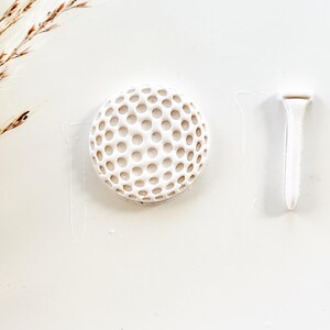 Pu&ograve; includere: Una pallina da golf bianca e un tee su sfondo bianco. La pallina da golf ha una superficie a fossette. Il tee ha una forma semplice e allungata. L'immagine &egrave; una vista dall'alto, con ombre sottili.