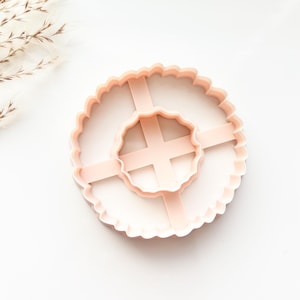 May include: A light pink, scalloped-edge cookie cutter with a cross-shaped interior design. The cutter is made of plastic and is designed for baking. The image is taken from a top-down perspective, with a white background.
