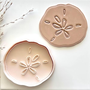 May include: Two sand dollar shaped cookie cutters, one is a solid pink color and the other is a white outline with a pink interior. Both cutters feature a detailed design of a sand dollar with a starburst pattern in the center.