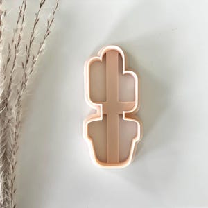 May include: A light pink cactus-shaped cookie cutter. The cutter is made of plastic and has a detailed outline of a cactus in a pot. The design is suitable for making cookies or other baked goods with a desert theme.