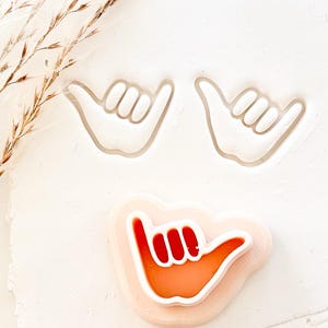May include: Three cookie cutters in the shape of the shaka hand gesture. Two are outlines, and one is filled with orange and red. The cutters are on a white surface.