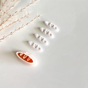 May include: An overhead shot shows a collection of miniature bread-shaped items. Four white, loaf-shaped pieces are arranged diagonally, with a fifth, orange-filled bread-shaped item at the bottom left. The items are on a white surface, with a few sprigs of dried grass in the background.
