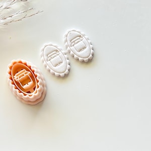May include: A light peach-colored cookie cutter with a scalloped edge, next to two cookies with the same design. The cutter is oval-shaped and features a detailed design of a jar. The cookies are white and have the same jar design.