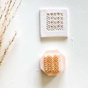 May include: Two craft stamps. One is a white square with a carved chevron pattern. The other is a light pink octagon with a matching chevron design. The stamps are for crafting and creating patterns.