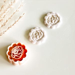 May include: Three rose-shaped cookie cutters. One is a vibrant red-orange color, while the other two are white. The cutters have a scalloped edge and a detailed rose design in the center. The background is a clean, white surface.