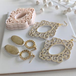 May include: A pair of gold and white statement earrings with a filigree design. The earrings are made of polymer clay and have a gold chain and bead detail.