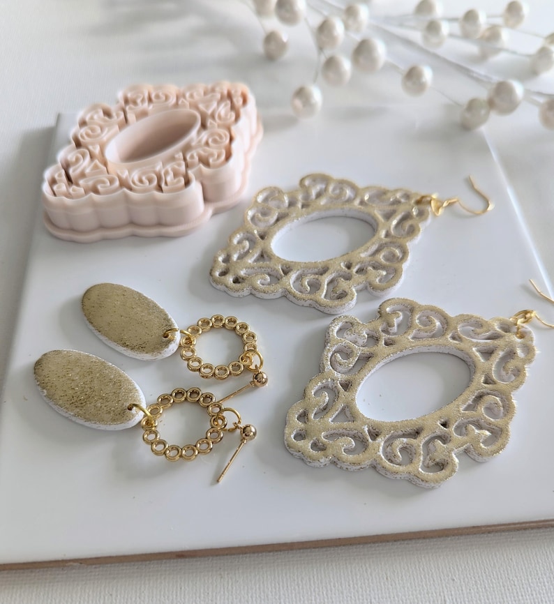 May include: A pair of gold and white statement earrings with a filigree design. The earrings are made of polymer clay and have a gold chain and bead detail.