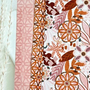 May include: A close-up of patterned fabric with floral and geometric designs. The color palette includes shades of orange, pink, and white. The fabric features large flower motifs, leaves, and geometric borders, creating a vibrant and detailed textile.