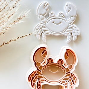 May include: Two crab-shaped cookie cutters. One is white, and the other is a light brown color. The crab cutters have a smiling face and detailed claws and legs. The cutters are on a white surface.