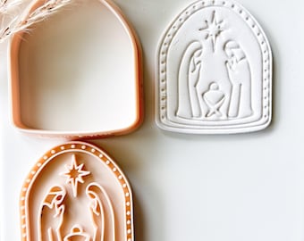 Könnte beinhalten: Ein pfirsichfarbener Kunststoff-Ausstecher und ein passender Stempel mit einer bogenförmigen Krippenszene, die zwei Figuren, ein zentrales Kind und einen Stern darüber zeigt. Der Stempel erzeugt einen erhabenen Abdruck in Teig oder Ton.