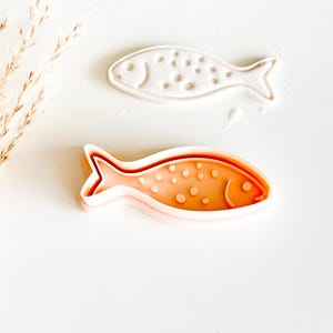 May include: Two fish-shaped cookie cutters. One is white, the other is orange. Both have a dotted pattern and a smiling face. The cutters are likely used for baking or crafting, adding a whimsical touch to treats or projects.