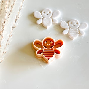 May include: Three bee-shaped items are displayed on a white surface. Two are white, formed from a soft material, and the third is a peach-coloured plastic cookie cutter. The bees have wings, smiling faces, and striped bodies.
