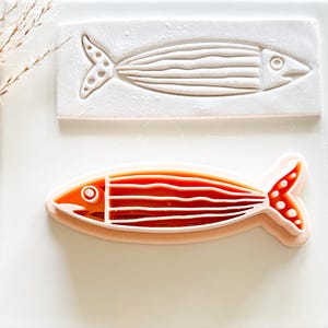 May include: A fish-shaped ceramic mold and a finished cookie cutter. The cookie cutter is orange and white, with a fish design. The mold is white, with a fish design. The fish has a tail, eye, and horizontal lines.