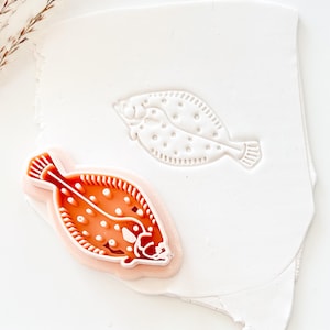 Peut inclure: Un emporte-pièce en forme de poisson rouge et blanc avec un design détaillé, à côté d'une empreinte blanche de la même forme. Le découpoir a une bordure rose et est sur une surface blanche. Le design comprend des points et des nageoires.