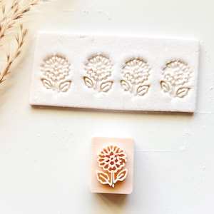 May include: A white rectangular mold with four flower impressions and a stamped flower design on a pink rectangular block. The flower design features a detailed floral pattern with petals and leaves, ideal for crafting or decorative purposes.